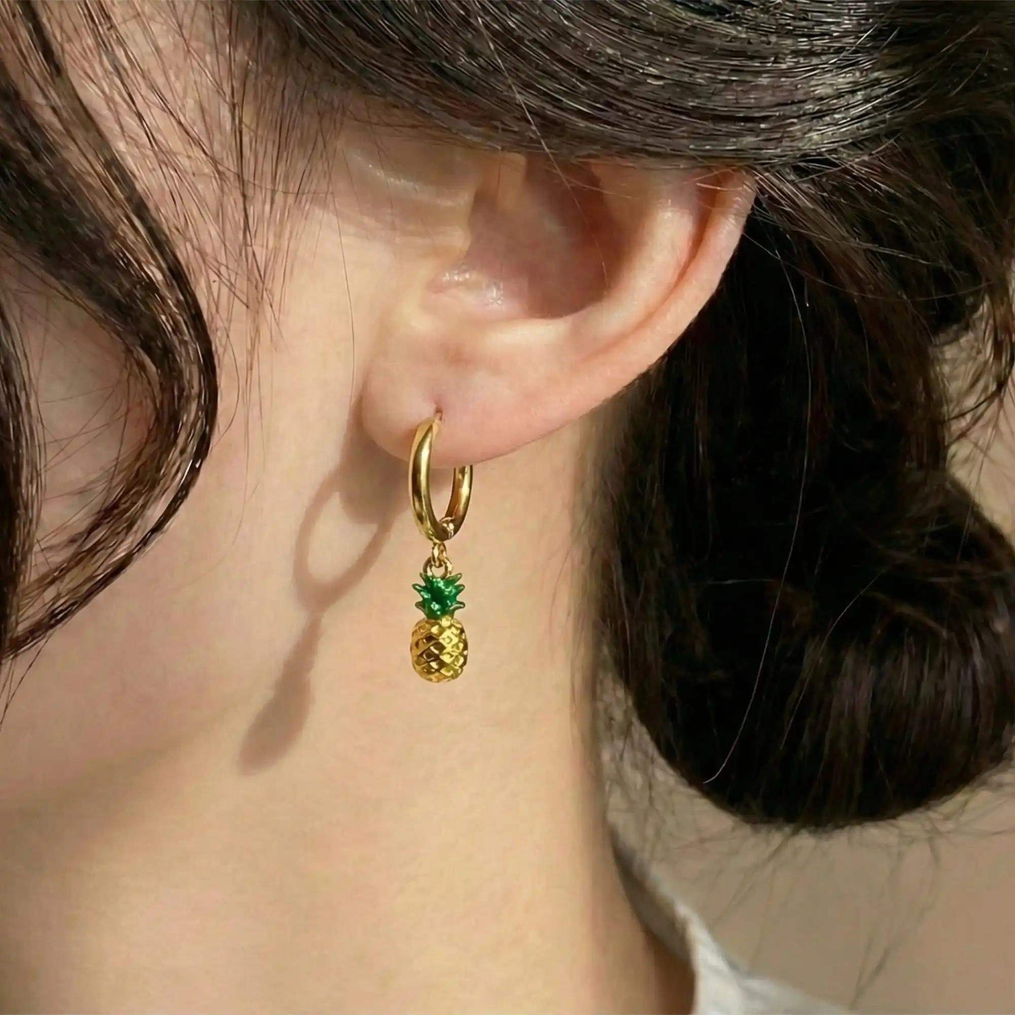 Close-up of a woman wearing a small gold hoop earring with a dangling pineapple charm featuring green enamel leaves.