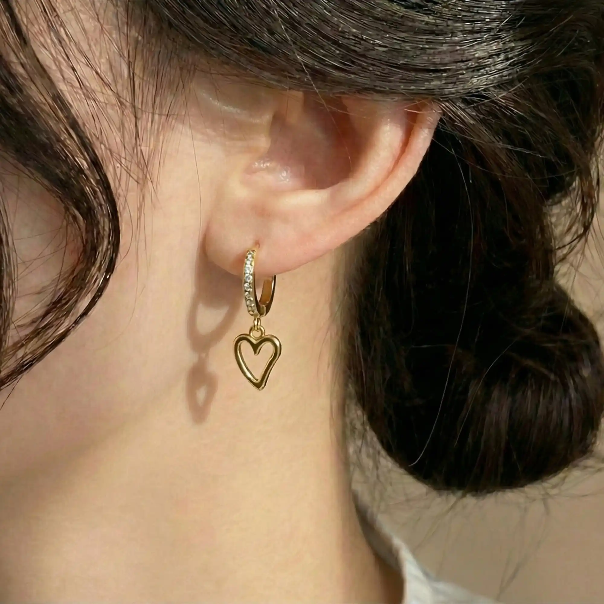 Close-up of a woman wearing a gold hoop earring lined with cubic zirconia stones, with a dangling open-heart charm